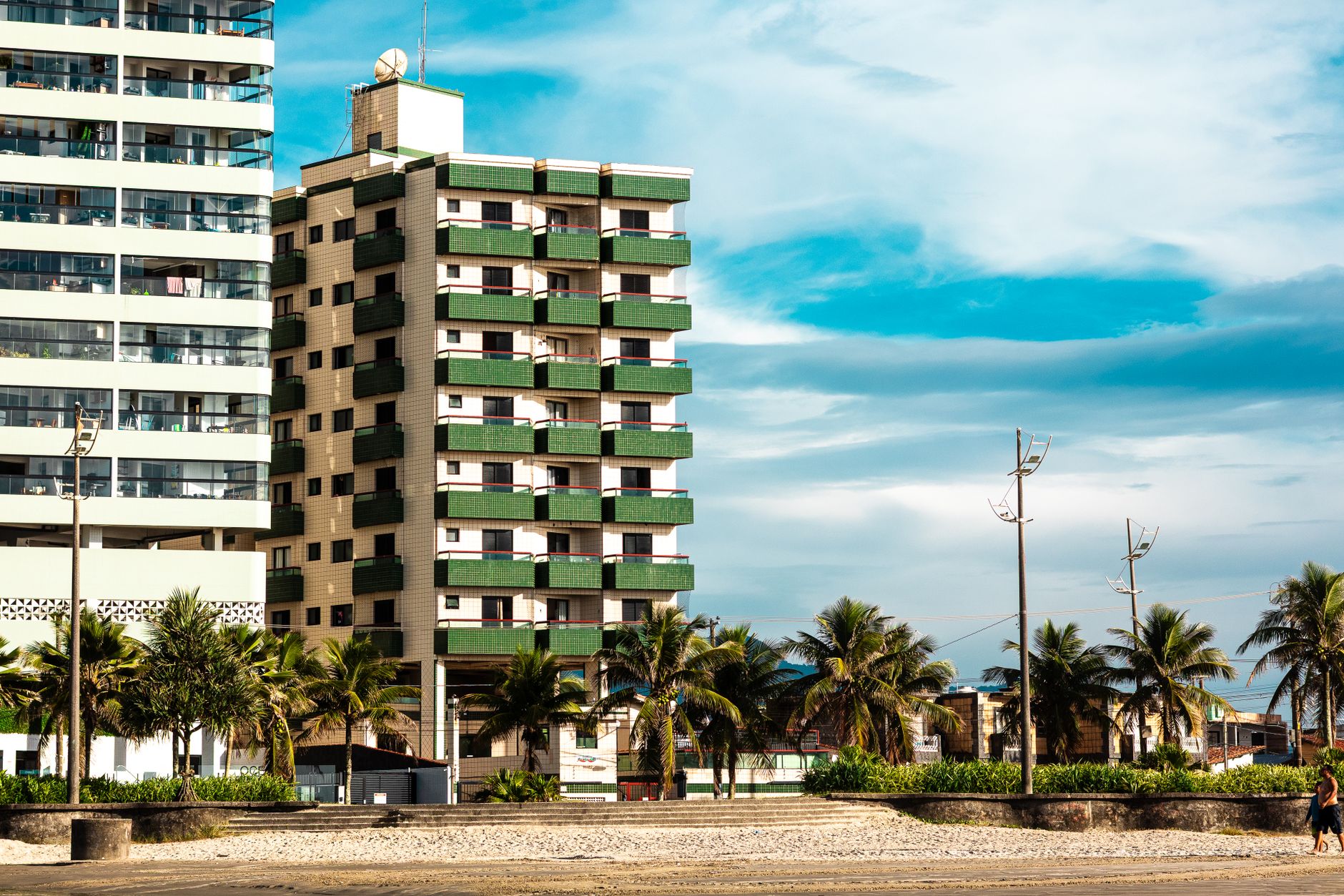 Um prédio de apartamentos moderno ao lado de uma praia tropical com palmeiras sob um céu azul límpido.