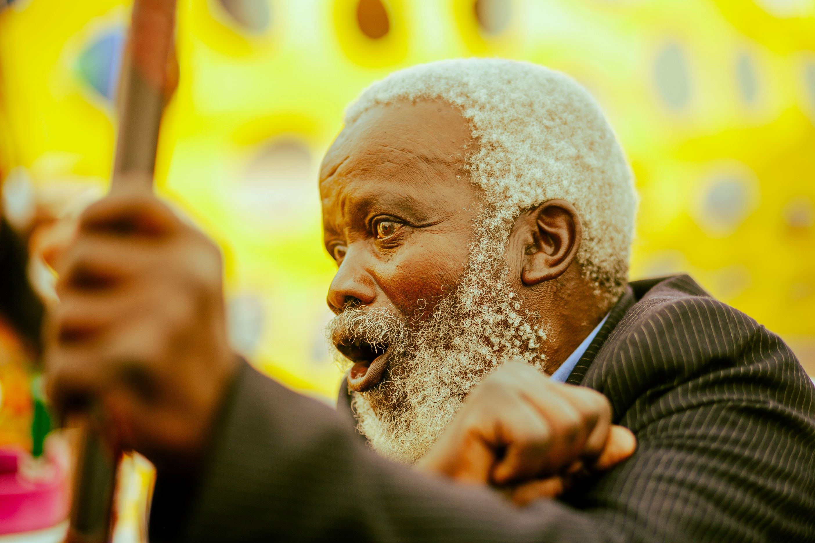 Dynamic portrait of a senior man with a white beard and expressive eyes against a bright yellow backdrop in Soweto.