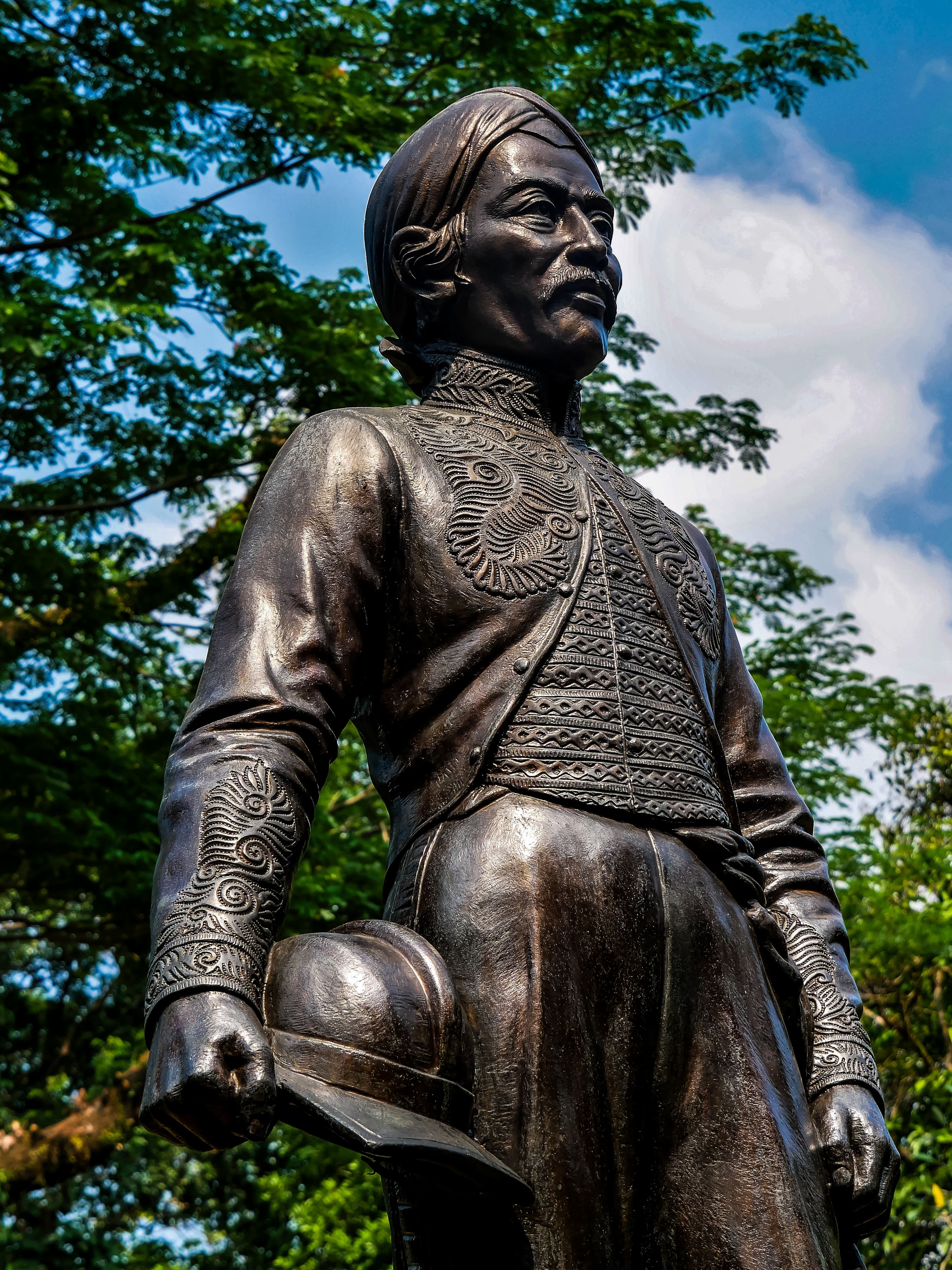 Bronze statue of historical figure with detailed attire, set against lush green foliage and blue sky.