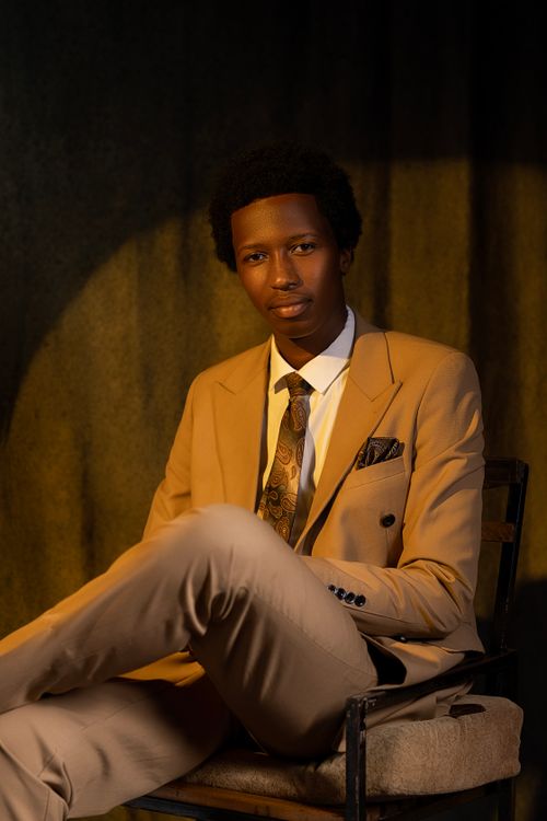 Free Young businessman in a brown suit sitting confidently indoors with warm lighting. Stock Photo