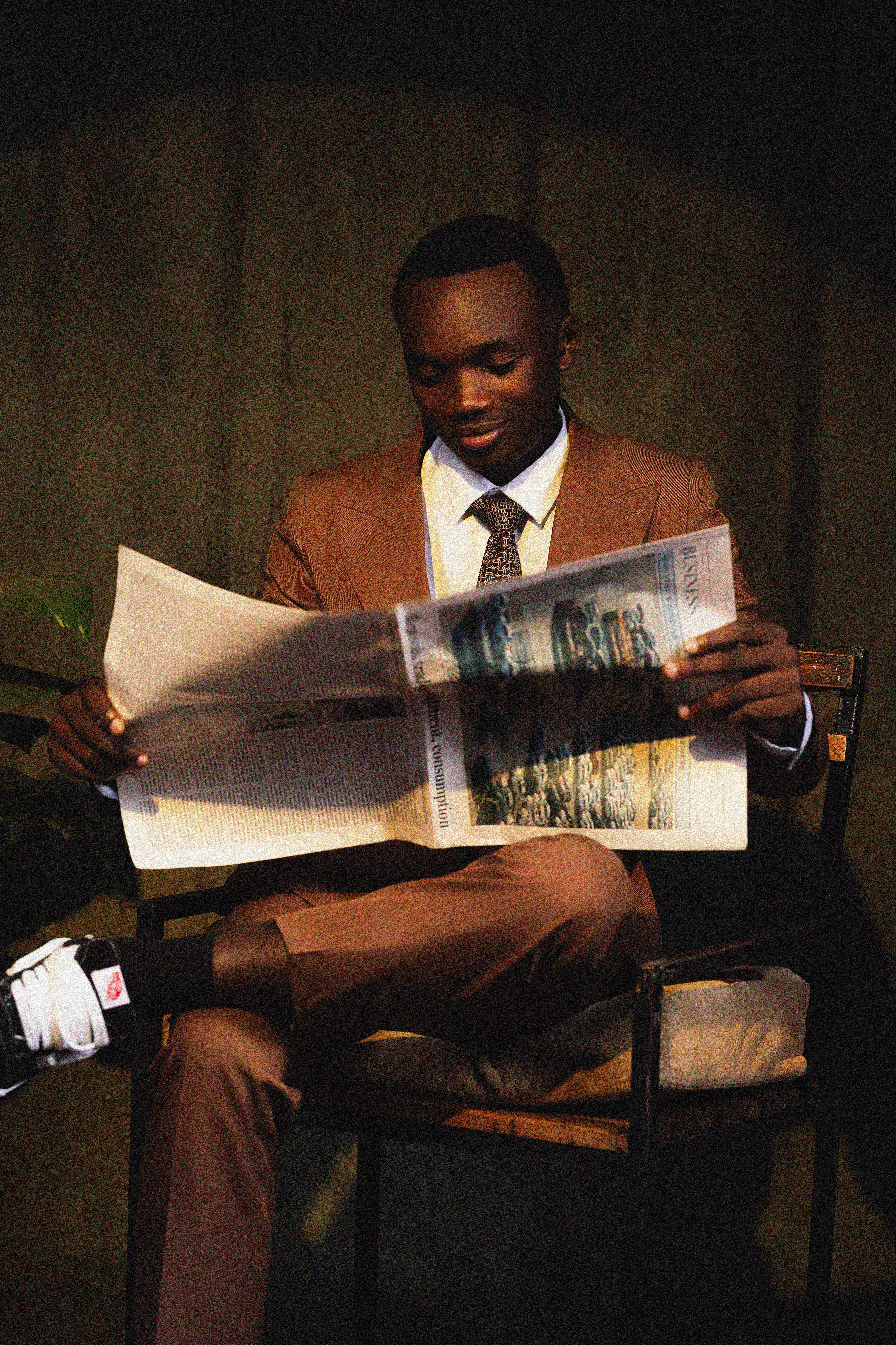 Well-dressed man in brown suit reading newspaper, seated indoors.