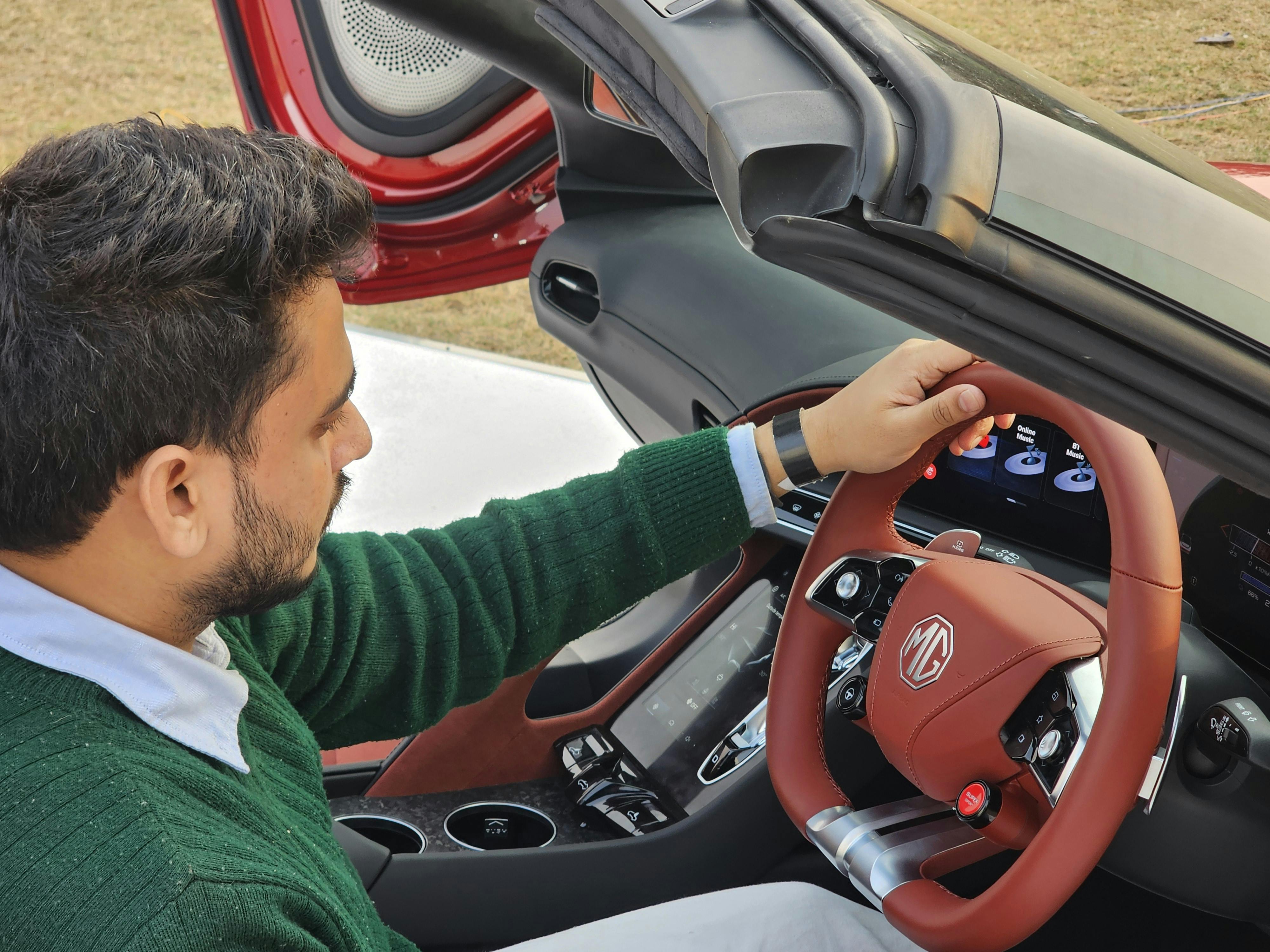 A man in a green sweater sits in a red convertible sports car, focusing on the controls.