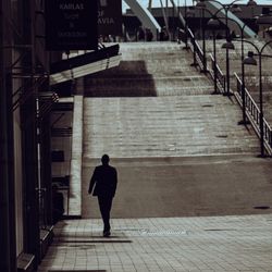 Une silhouette masculine marche sur une rue pavée en pente dans une zone urbaine, entourée de réverbères et de vitrines de magasins avec des enseignes, sous une lumière tamisée et dramatique.