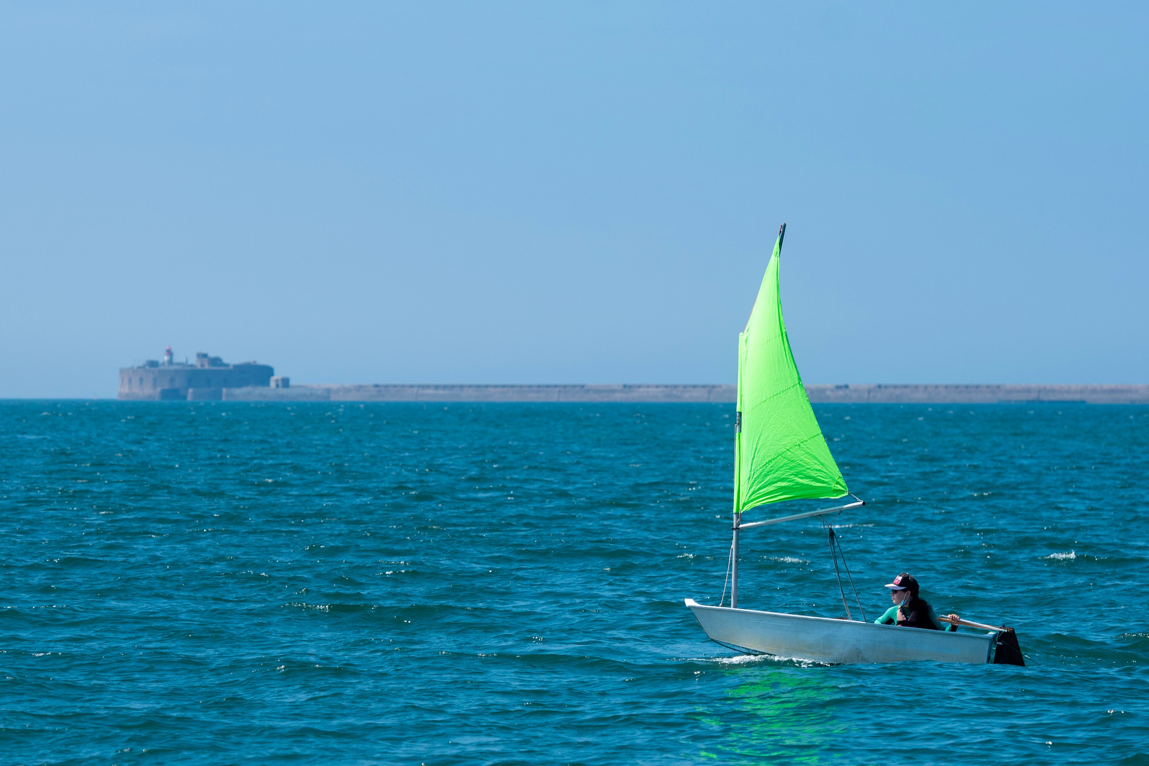 A person sailing a small boat with a bright green sail near Fort de Querqueville, Normandy.