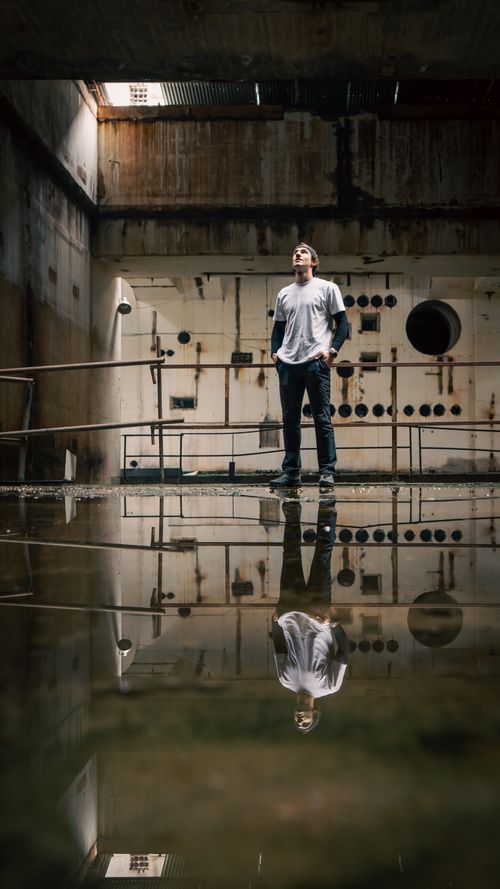 Free A man stands in a reflective puddle inside an abandoned industrial facility, creating an artistic scene. Stock Photo