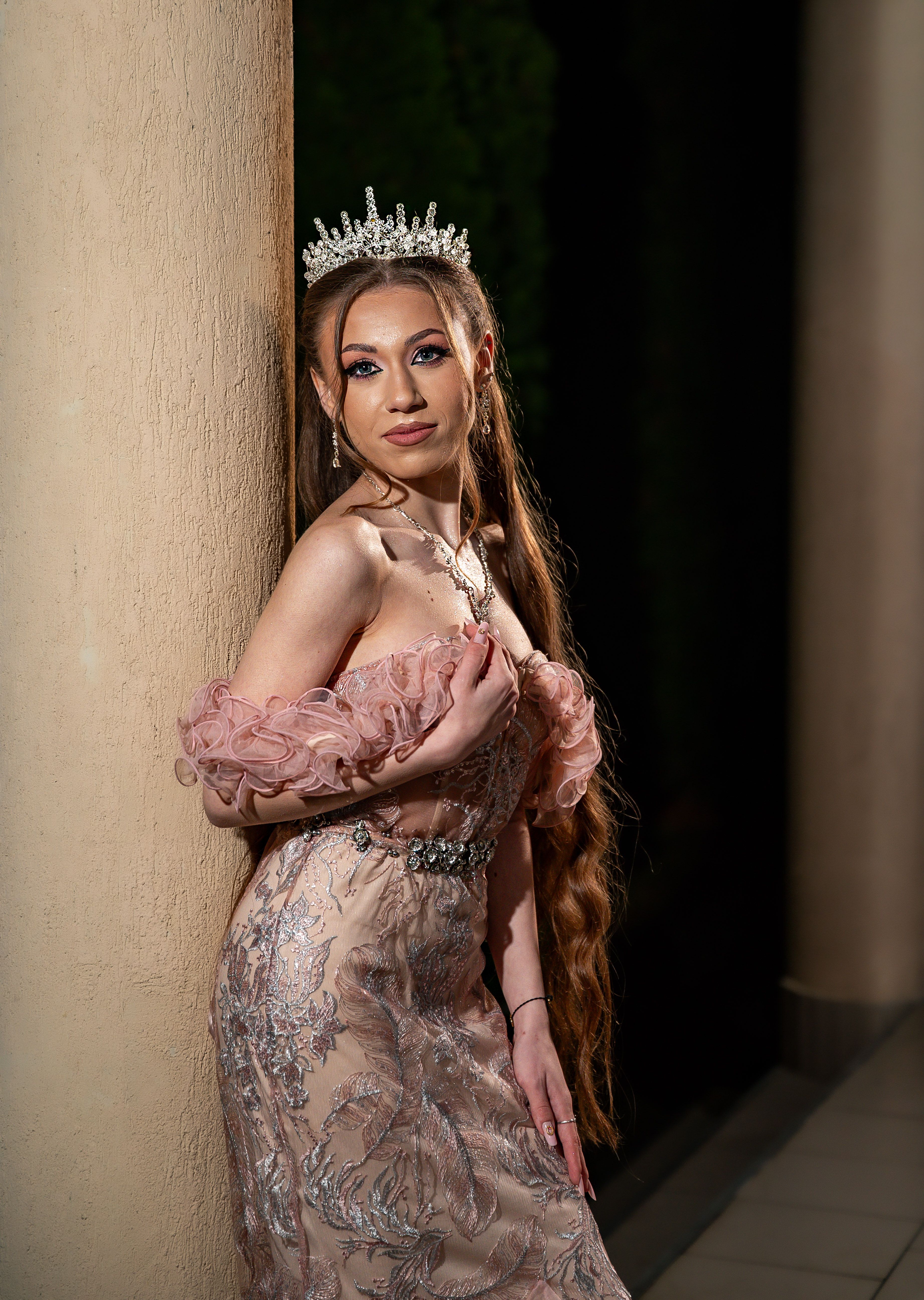 A beautiful woman in an ornate gown and tiara posing elegantly against a column.