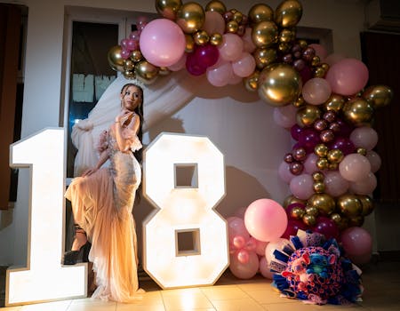 A young woman poses by illuminated 18, surrounded by colorful balloons in a festive indoor setting.