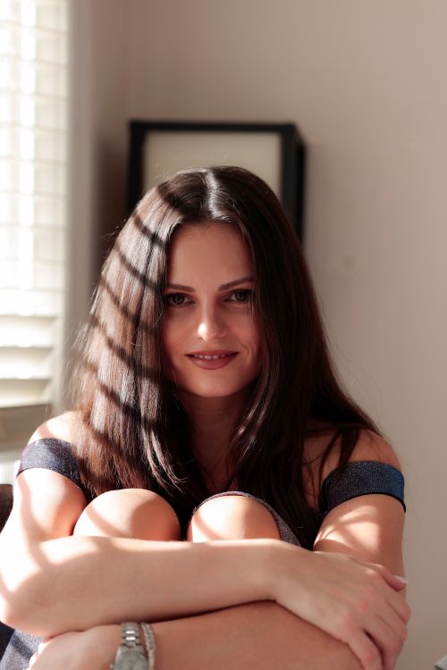 Free A woman sitting by a window, smiling gently, with sunlight casting shadows across her face. Stock Photo