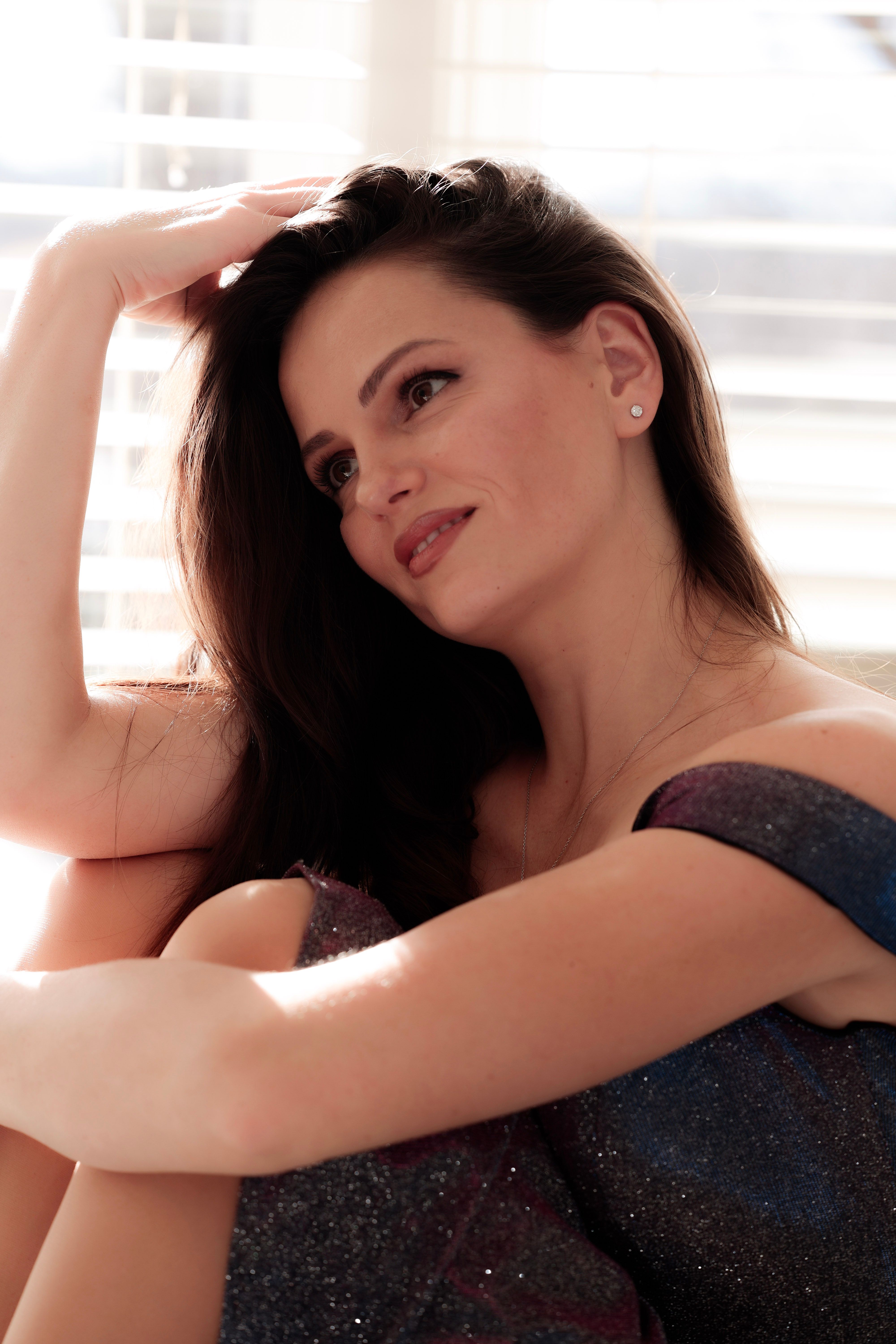 A serene and thoughtful woman in a sparkly dress sitting indoors with soft sunlight.