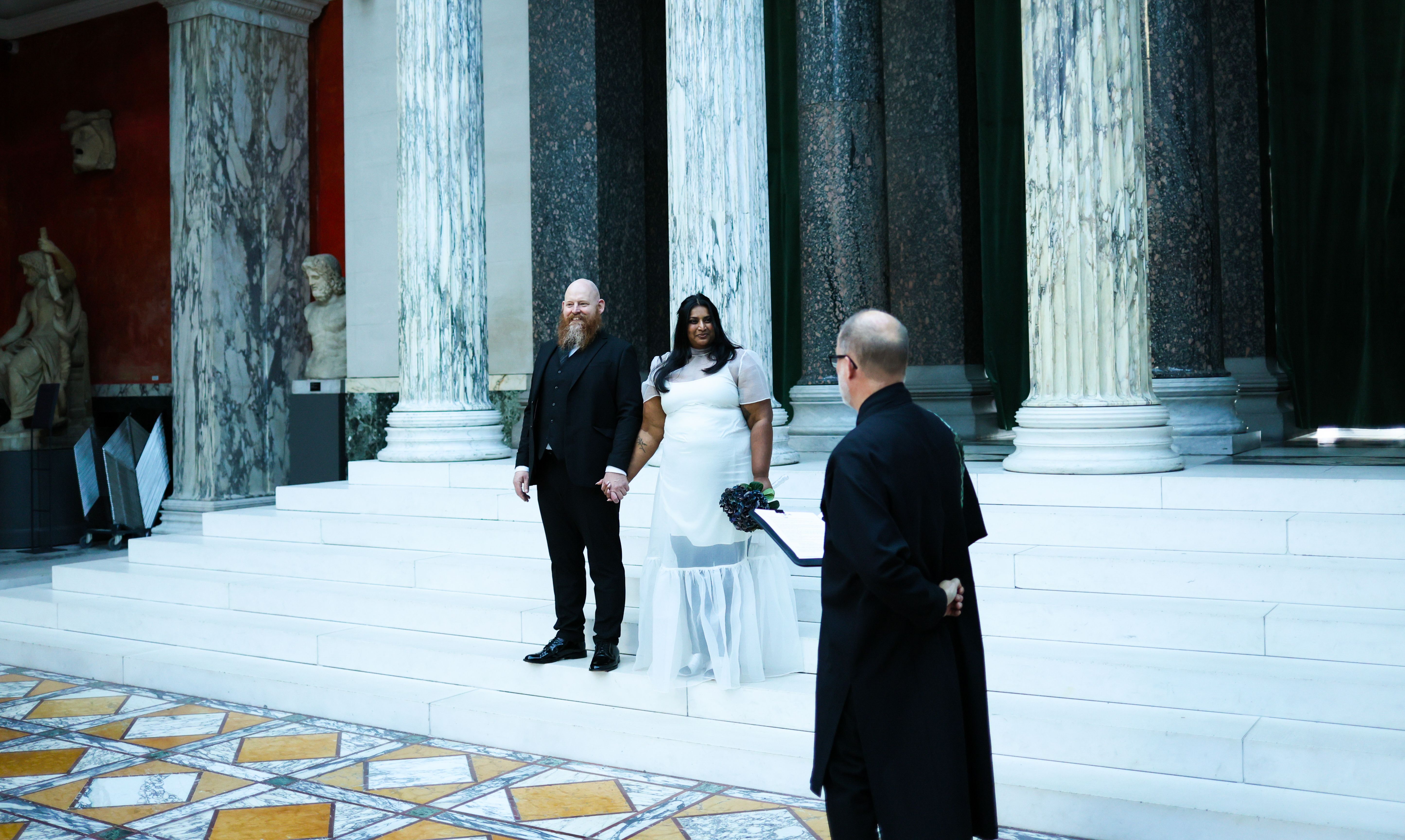 A couple stands together during their wedding ceremony in a grand hall with columned architecture.
