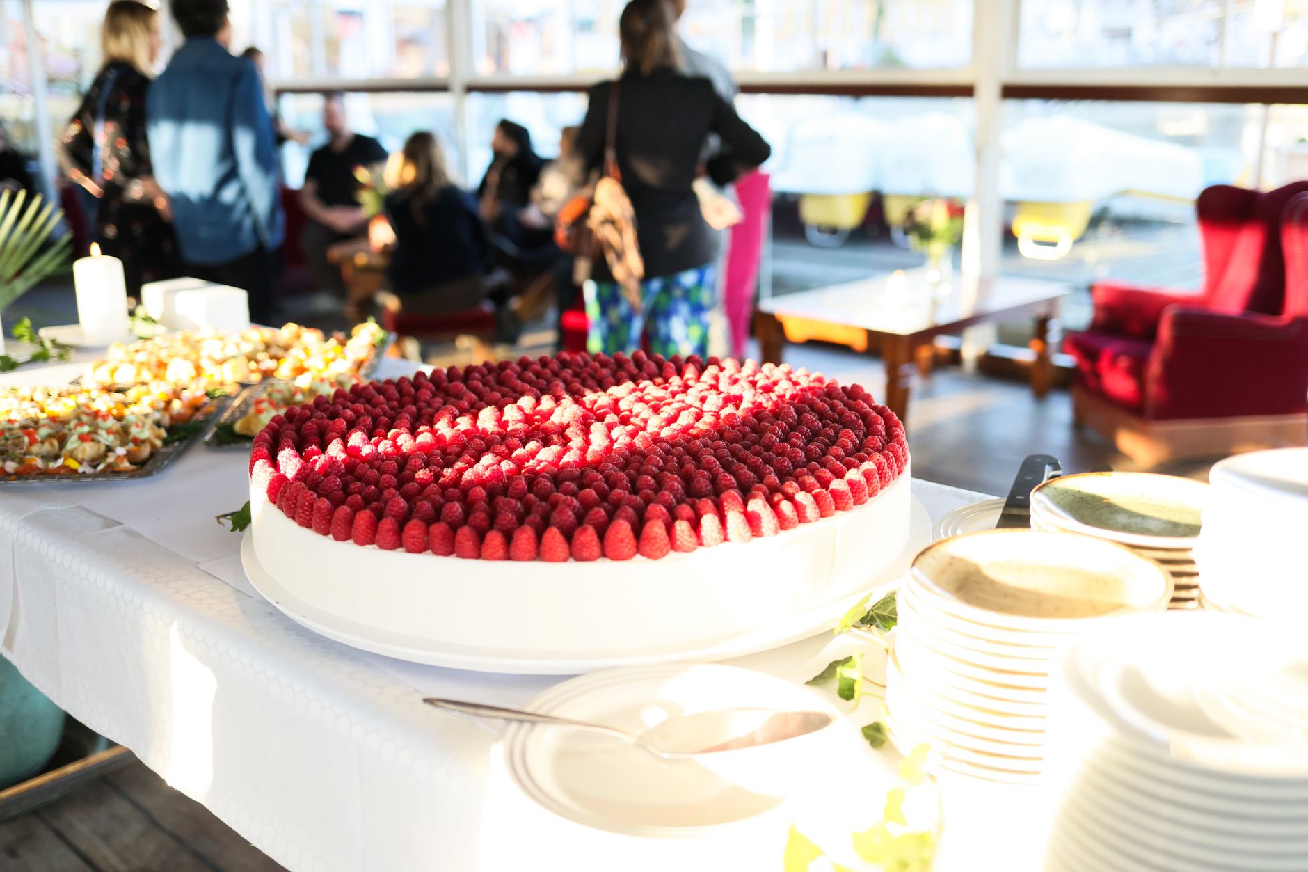 Raspberry covered cake on display at an indoor social event with diverse guests in the background.
