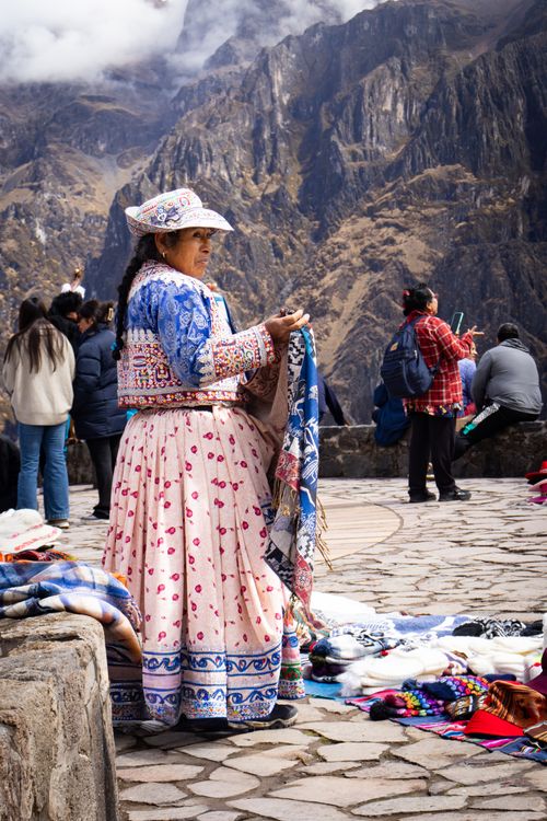 Free Peruvian woman in traditional attire selling handmade textiles at Colca Canyon, with Andes mountains as backdrop. Stock Photo