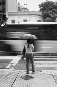 Black and white image of a woman standing at a city crosswalk with an umbrella as a bus speeds by.