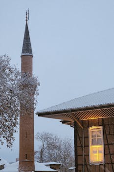 Minaret and building roof covered in snow during winter with lit window.