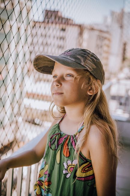 Young girl in casual summer dress and cap smiling outdoors against urban backdrop.