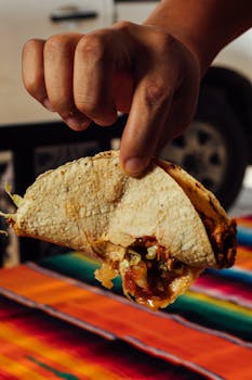 Enjoy a delicious taco held by a hand with vibrant Mexican textile in the background.