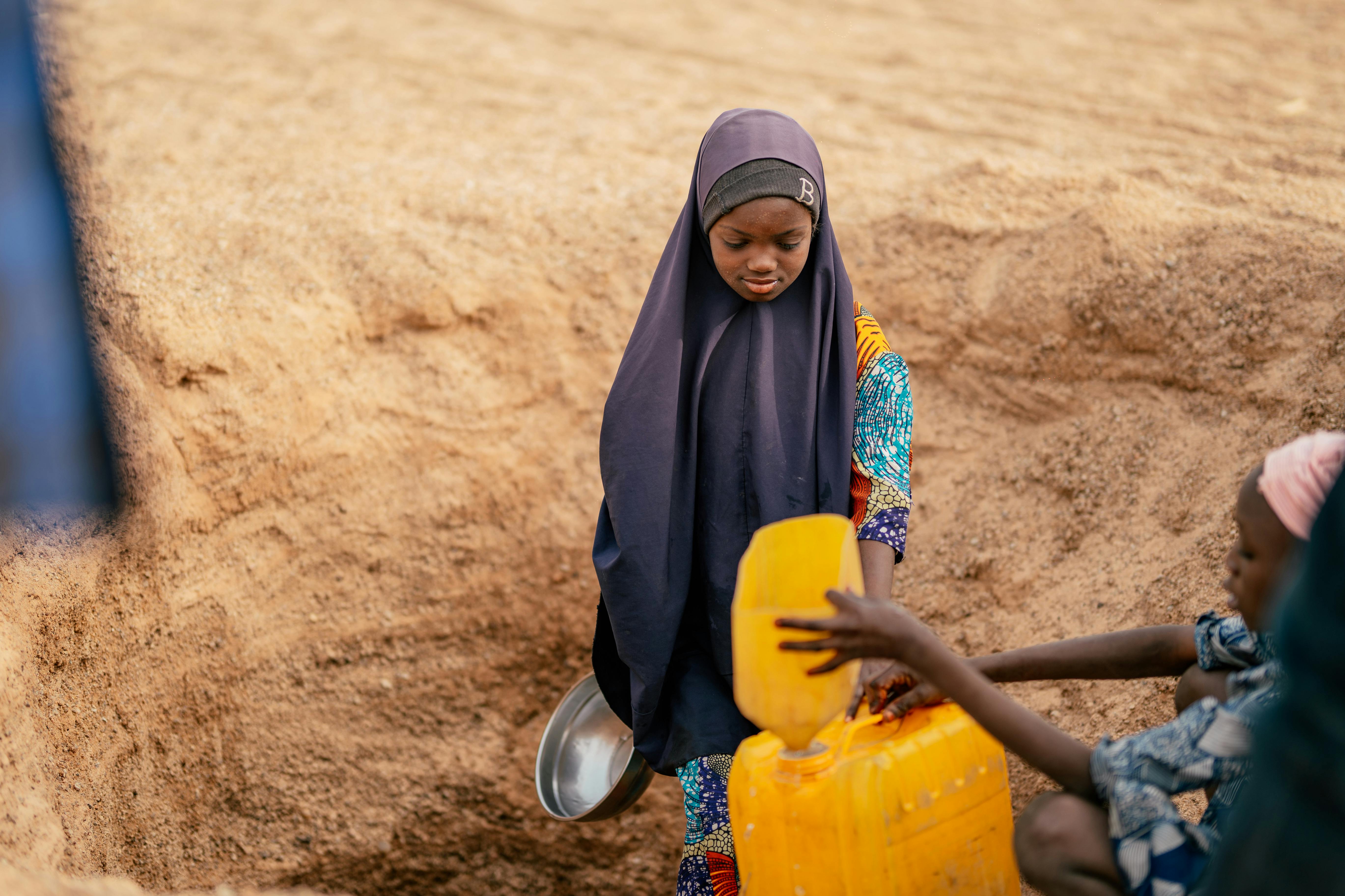A young girl in traditional attire gathers water from a sand pit in a rural setting.