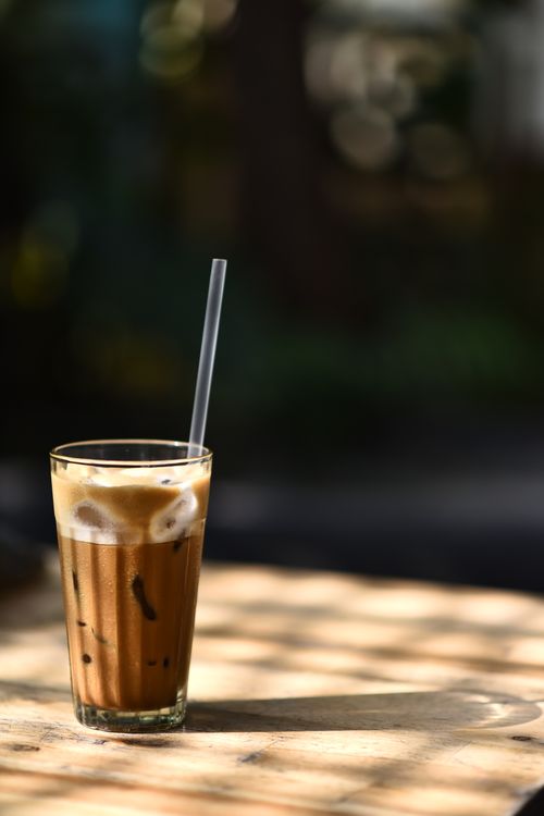Free A tall glass of iced coffee with a straw sits on a sunlit wooden table, perfect for a summer day. Stock Photo