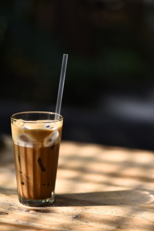 Free A tall glass of iced coffee with a straw sitting on a sunlit wooden table. Stock Photo