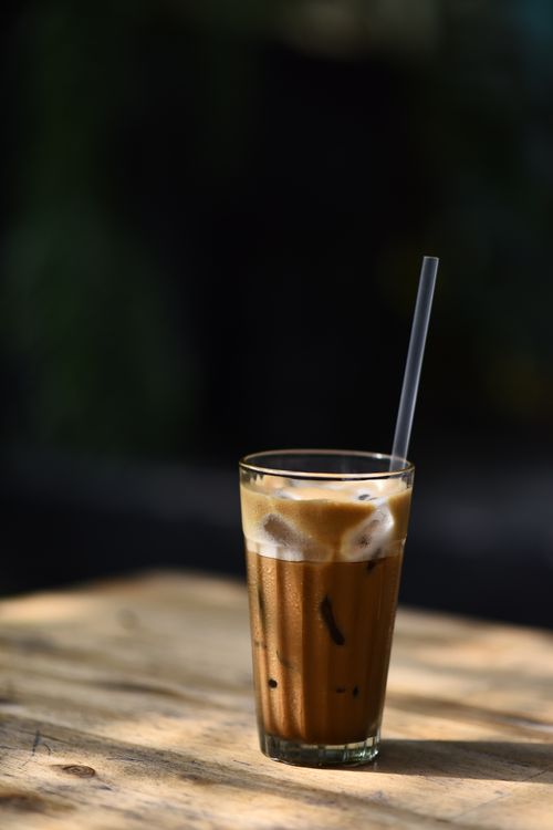 Free A tall glass of iced coffee with a straw, basking in warm sunlight on a wooden table. Stock Photo