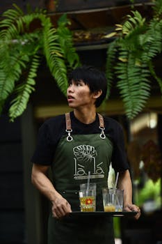 A young waiter in an apron carries drinks on a tray at an outdoor cafe under green ferns.
