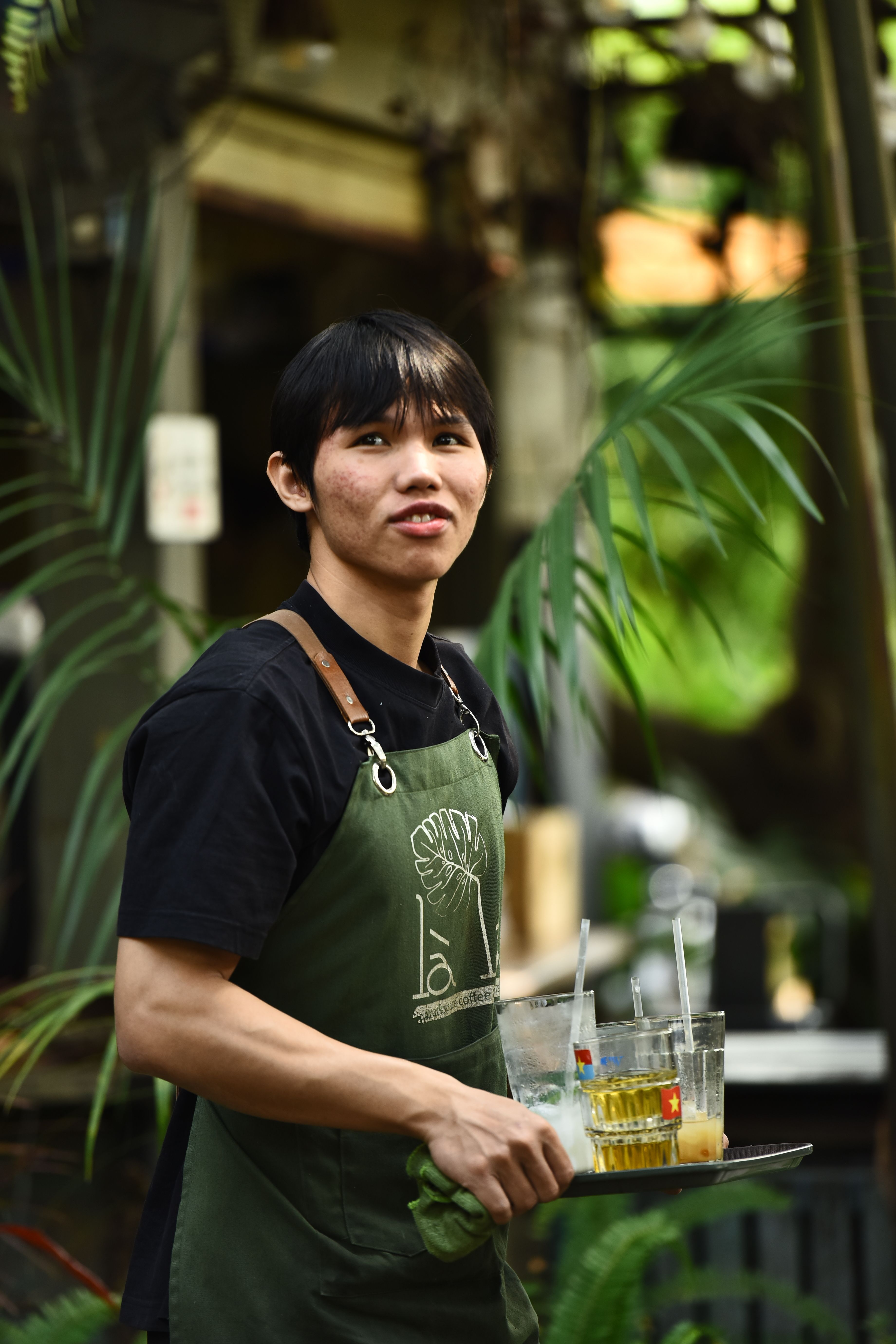 A young waiter holding drinks in an outdoor cafe setting, surrounded by plants.