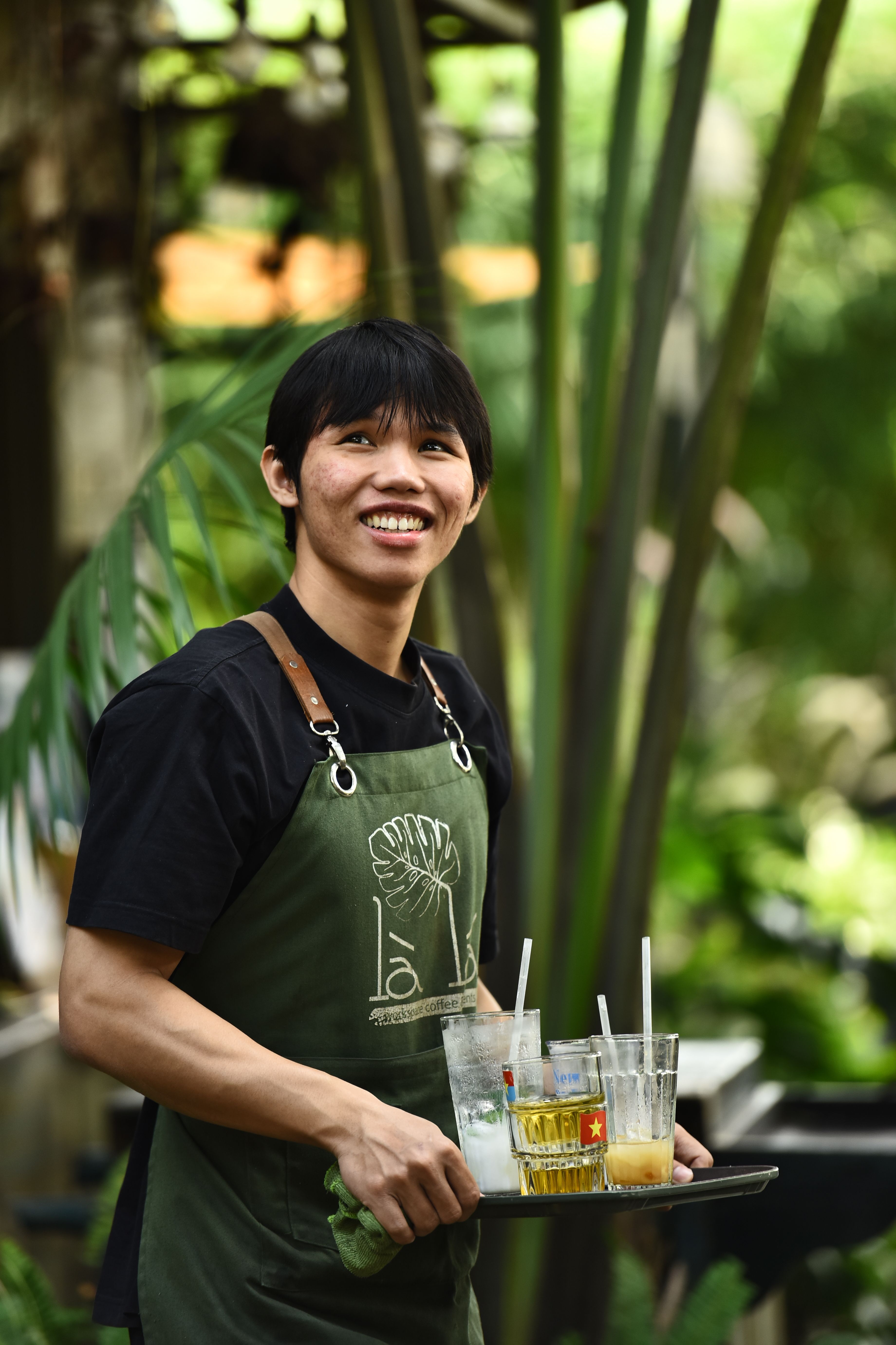 Friendly waiter outdoors holding a tray with colorful drinks, surrounded by lush greenery.