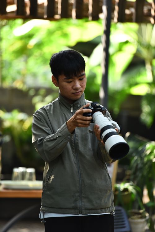 Free A young man in a green jacket adjusts his DSLR camera outdoors in a lush garden setting. Stock Photo
