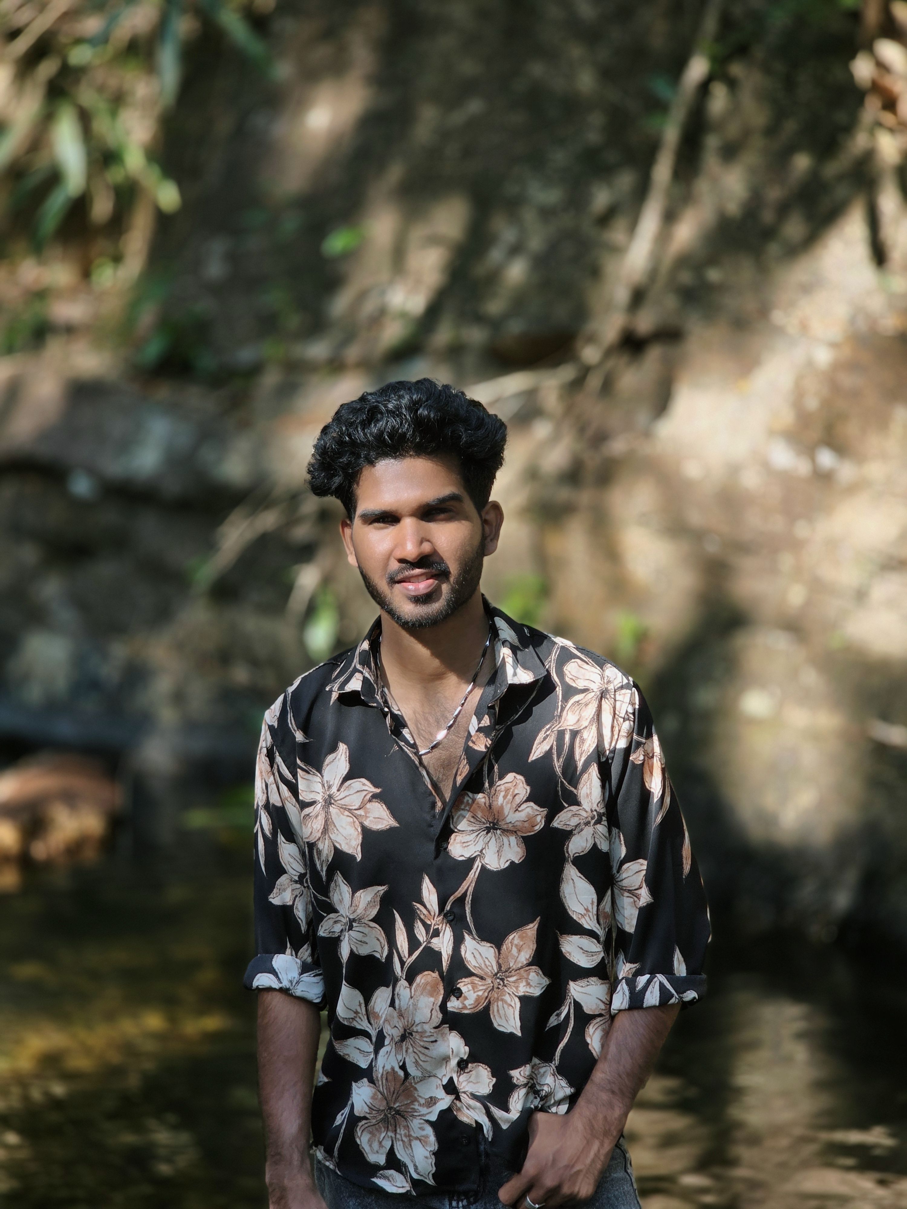 A young man in a floral shirt stands calmly by a forest stream.