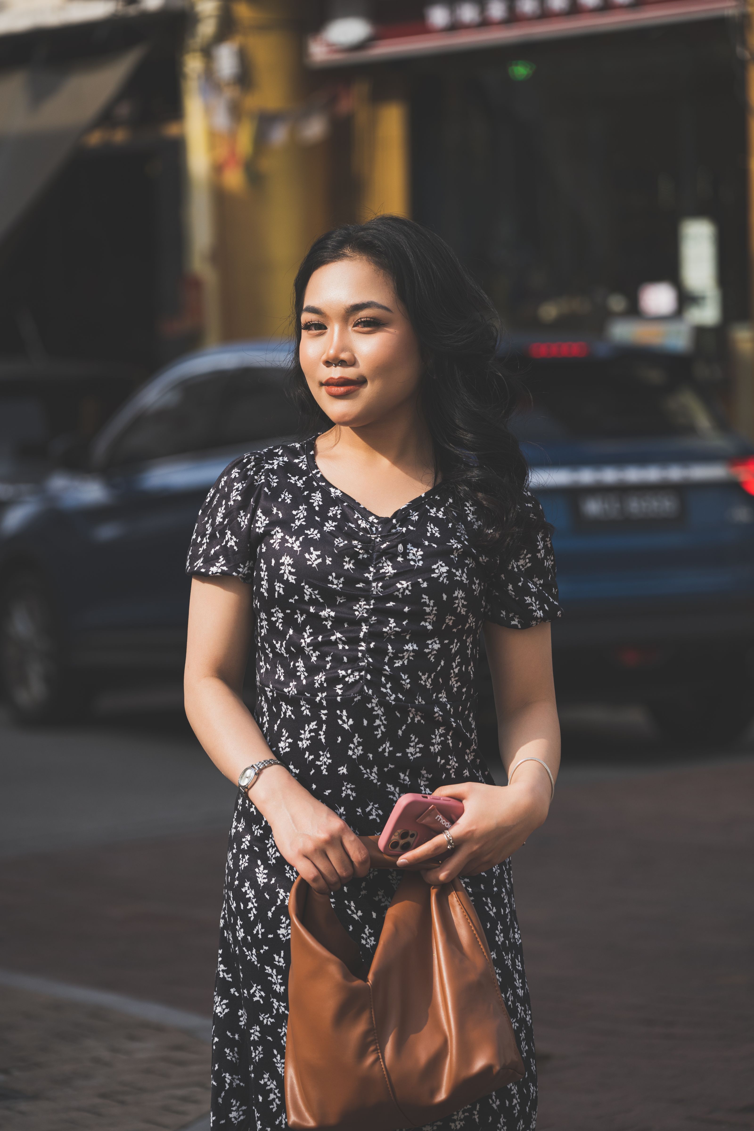 Jovem mulher de vestido florido, carregando uma bolsa, caminha pelo centro da cidade com carros ao fundo.