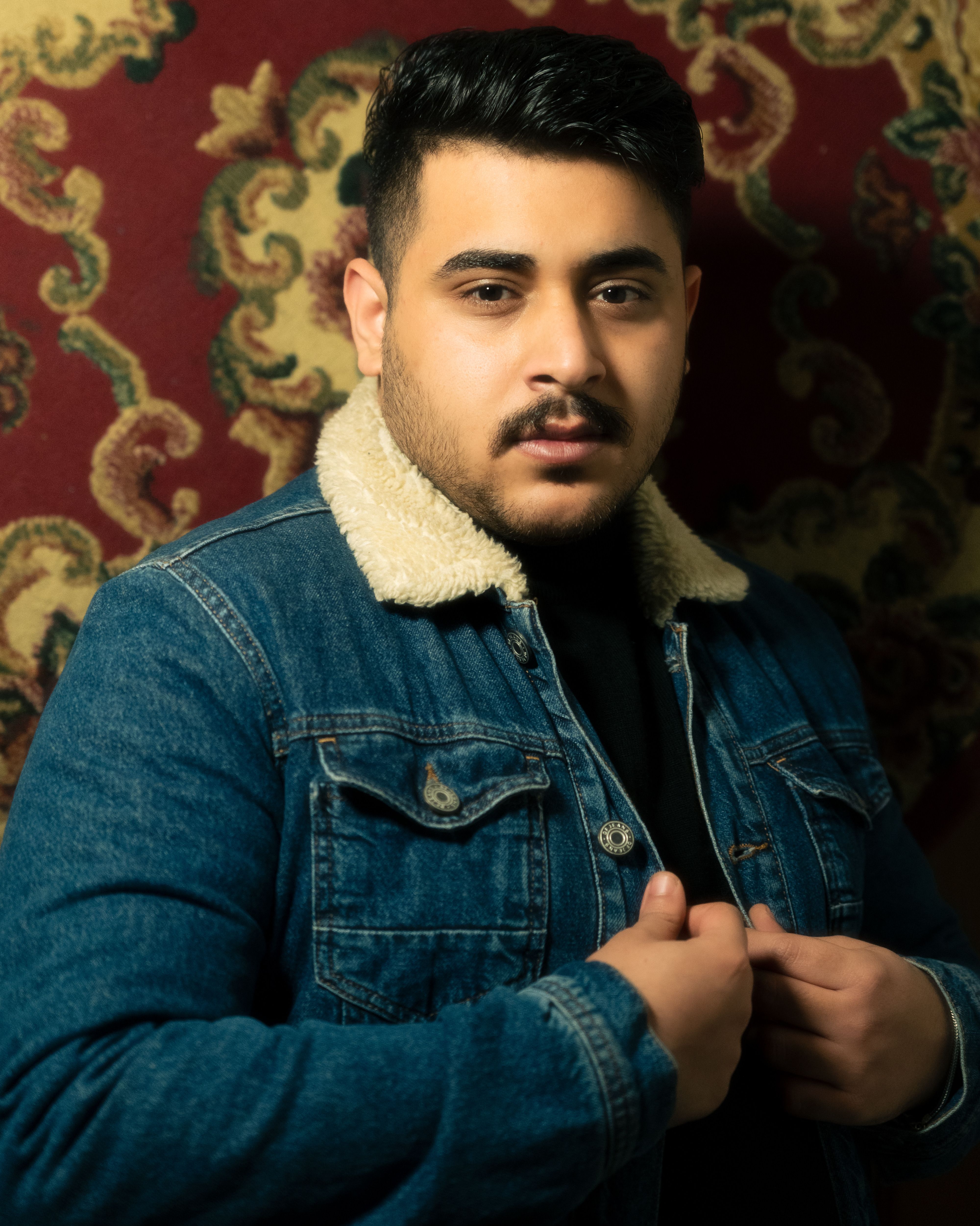 Confident young man in a denim jacket posing indoors with a patterned backdrop.
