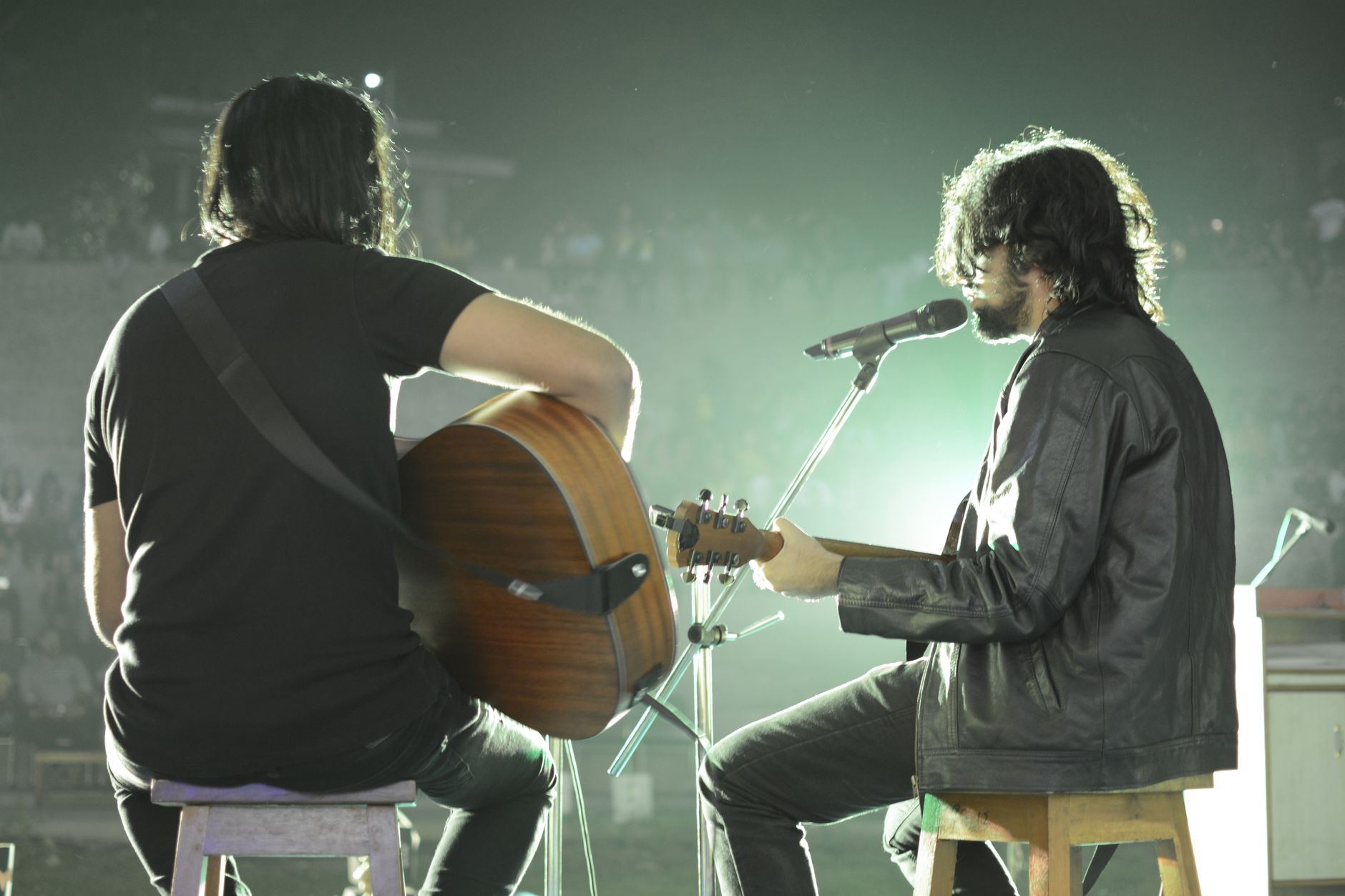 Men in casual clothes sitting on stool near microphone playing on guitars and singing while performing on stage during concert
