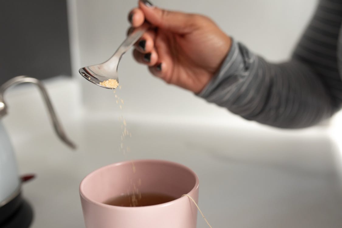 Unrecognizable female in casual clothes holding tea spoon and pouring sugar into ceramic cup with hot tea  on light kitchen