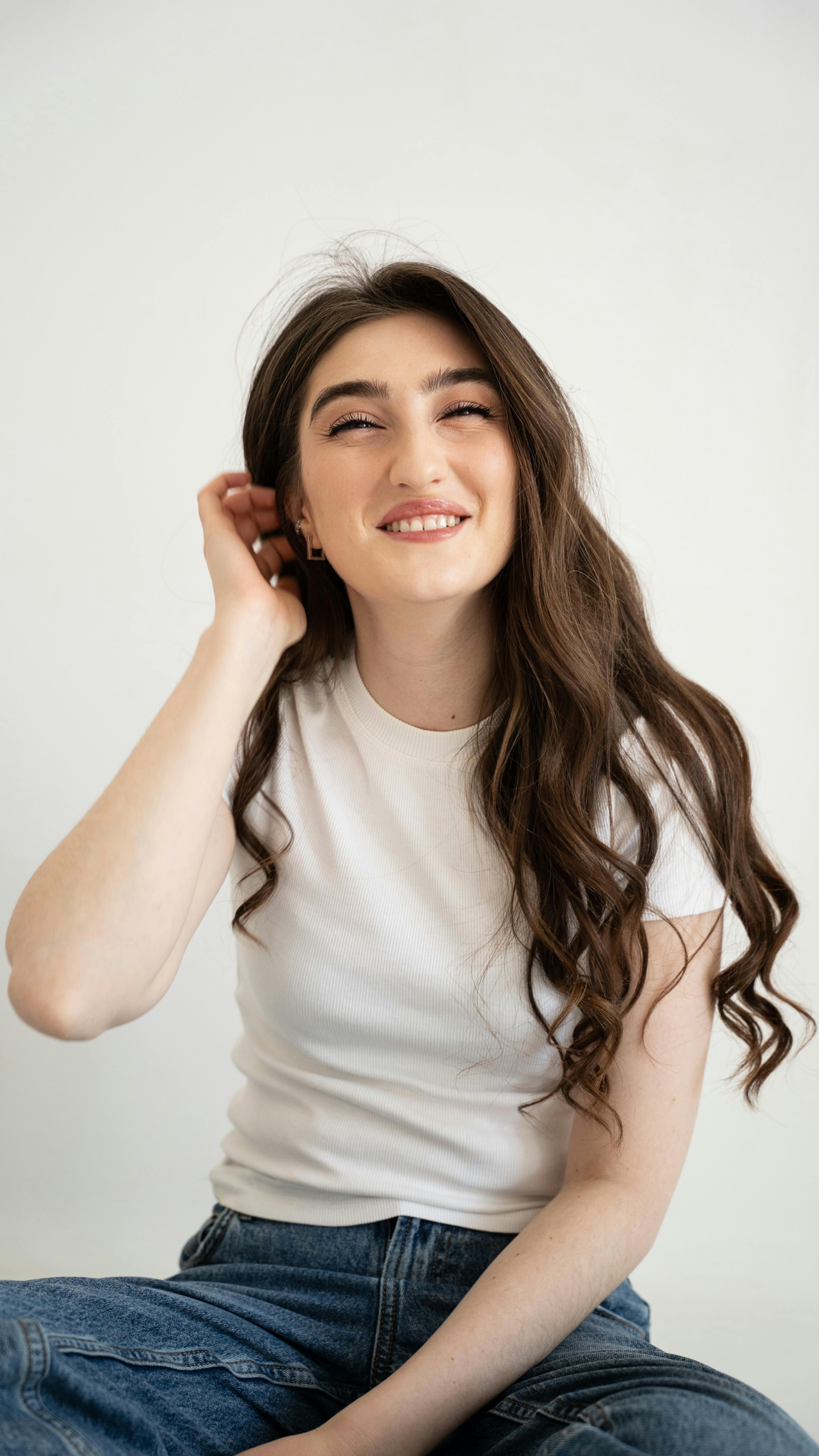 Miễn phí Chân dung một người phụ nữ mỉm cười, tóc dài, mặc áo sơ mi trắng và quần jeans, đang ngồi trong nhà. Ảnh lưu trữ