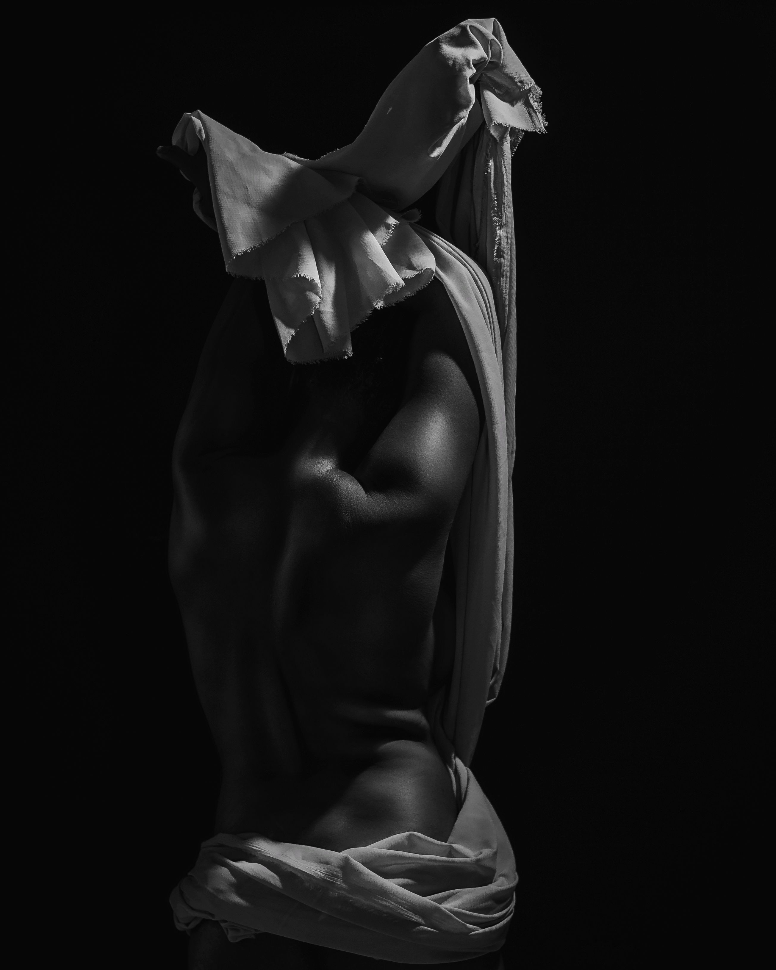 Black and white image of a nude figure in shadow, wrapped in cloth, highlighting form and structure.