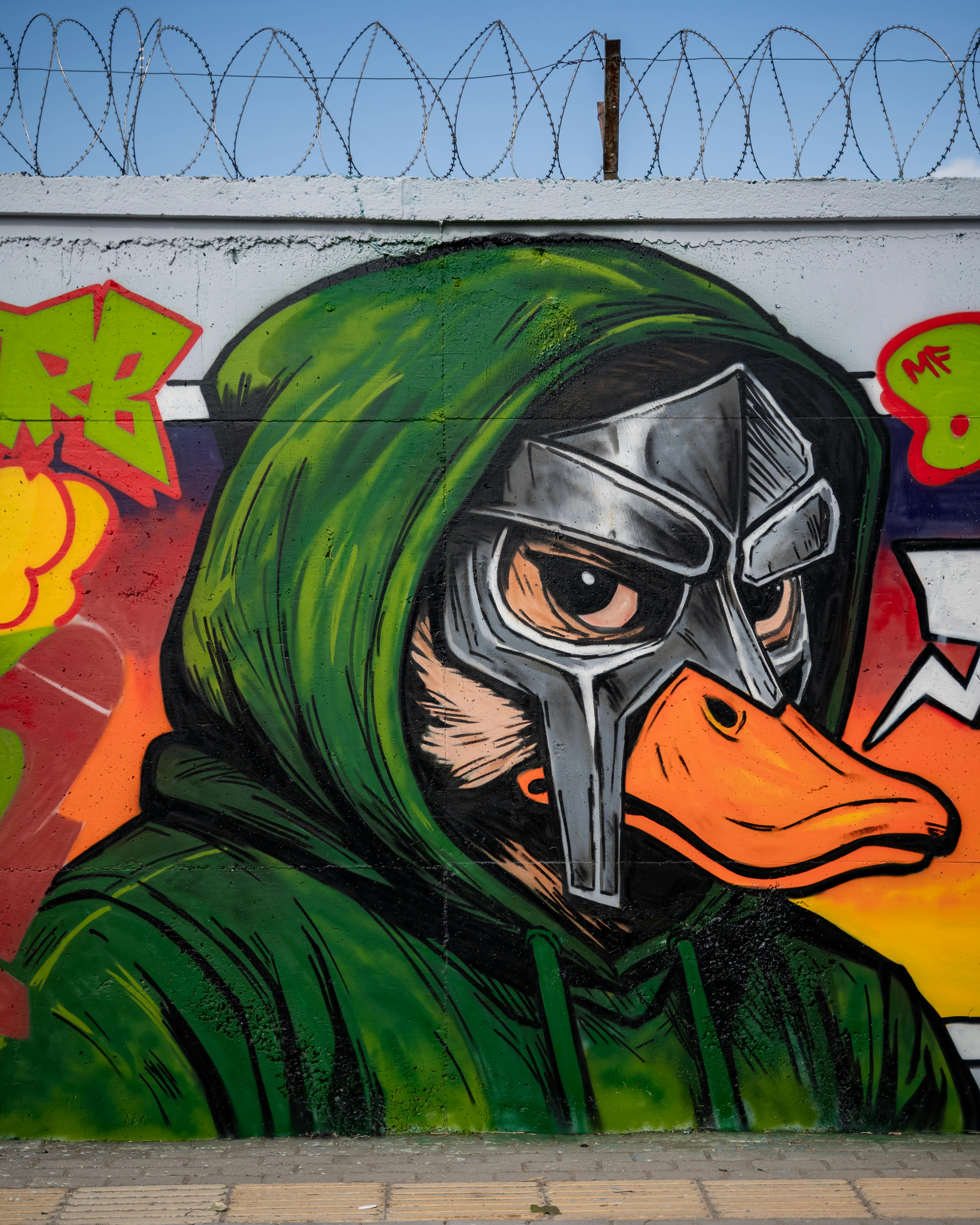 Vibrant mural of a duck in a hood and metal mask on a graffiti-covered wall.