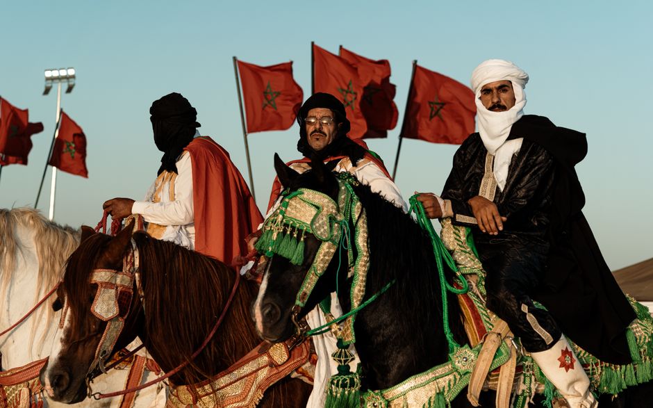 Traditional Moroccan horsemen in rich attire against a backdrop of Moroccan flags at dusk.