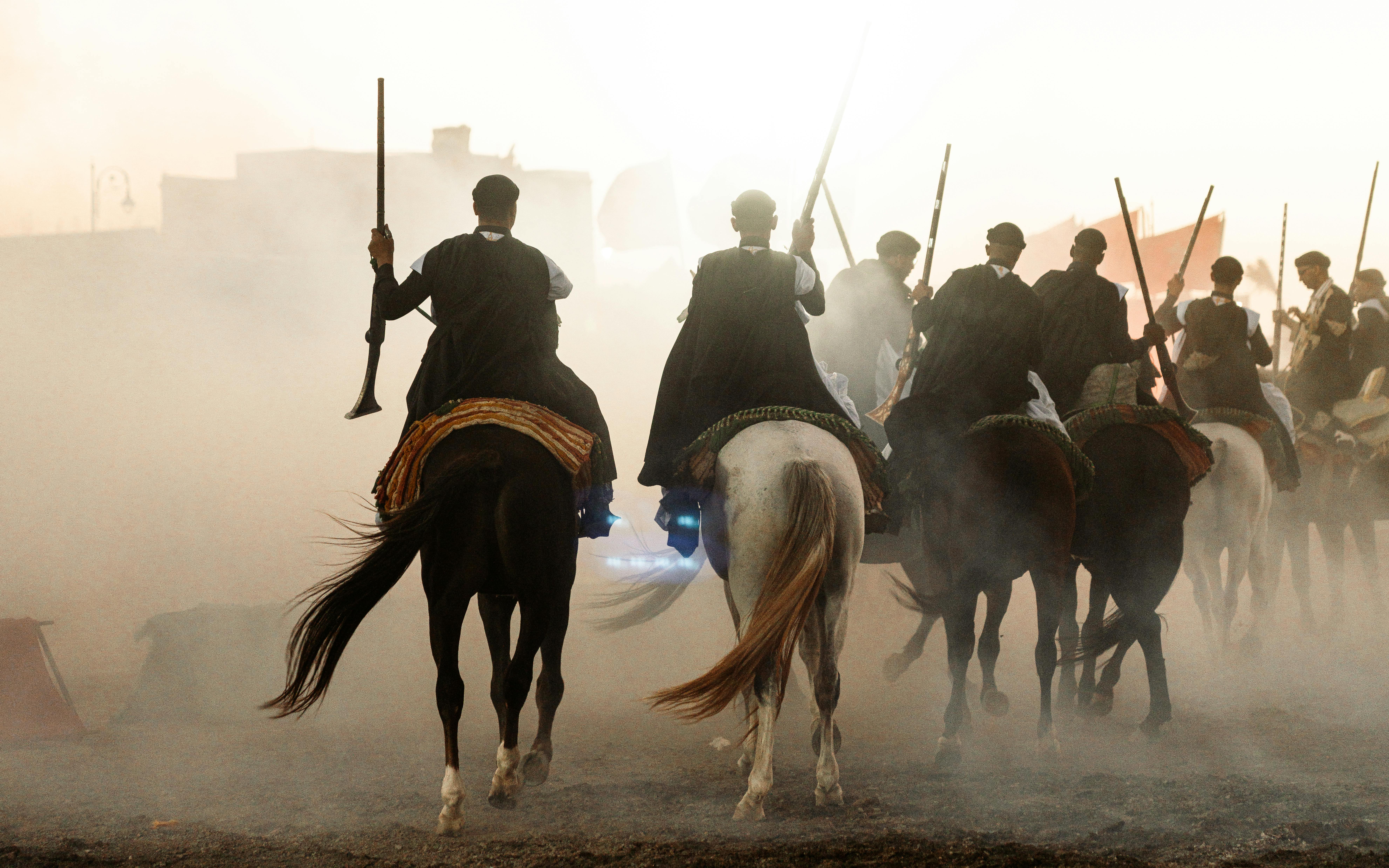 A group of horsemen perform the traditional Moroccan fantasia during sunset, creating dramatic dust clouds.