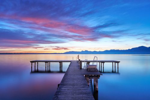 Free A tranquil view of Lake Chiemsee's pier at sunset with Alps in the background. Stock Photo