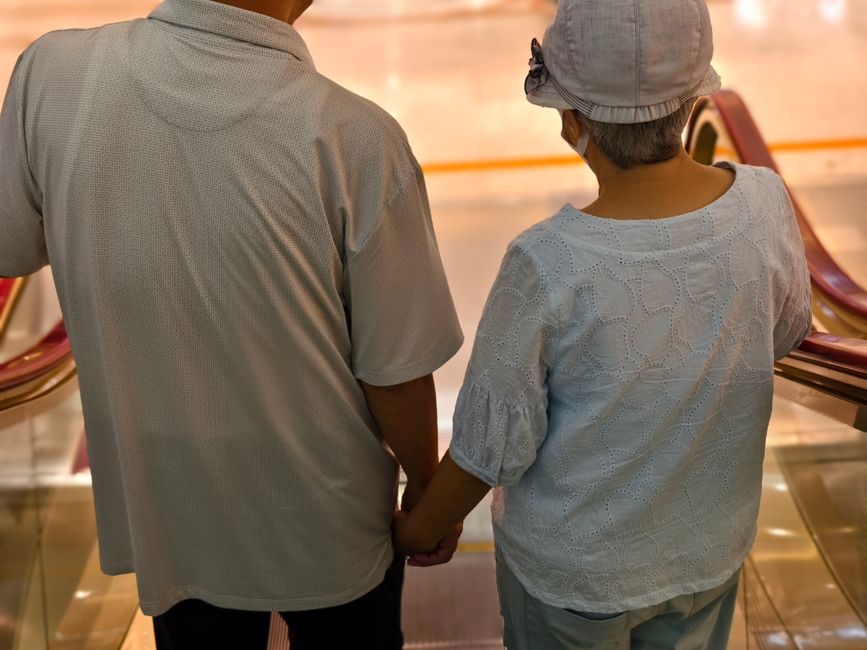 Elderly couple descends escalator hand in hand, symbolizing lasting love.