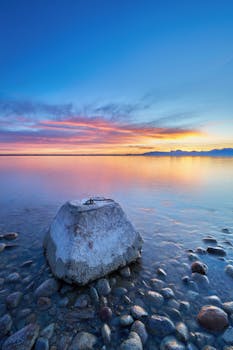 A tranquil sunrise over a calm Bavarian lake with colorful skies and rocky shore foreground.