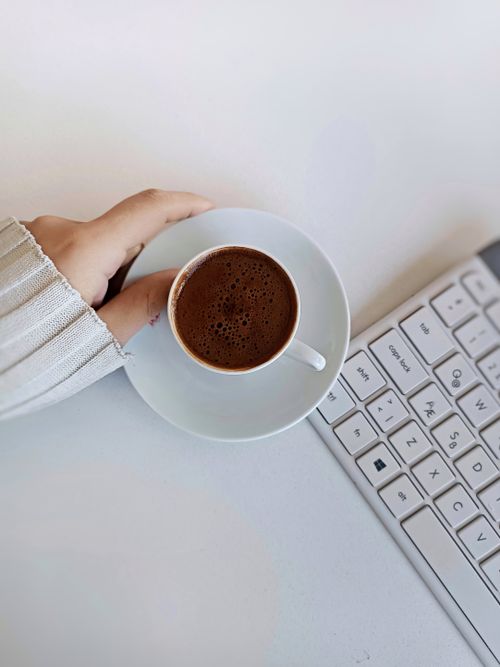 Free Hand holding a cup of coffee next to a white keyboard, creating a calm workspace vibe. Stock Photo