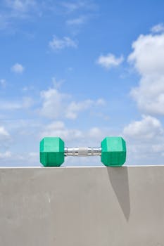 A green dumbbell rests on a beige wall under a clear blue sky, symbolizing fitness and wellness.