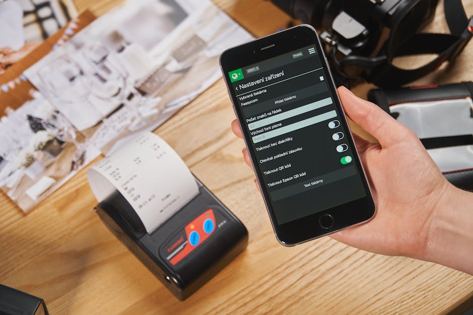 Close-up of a hand holding a smartphone with POS settings displayed, next to a receipt printer.