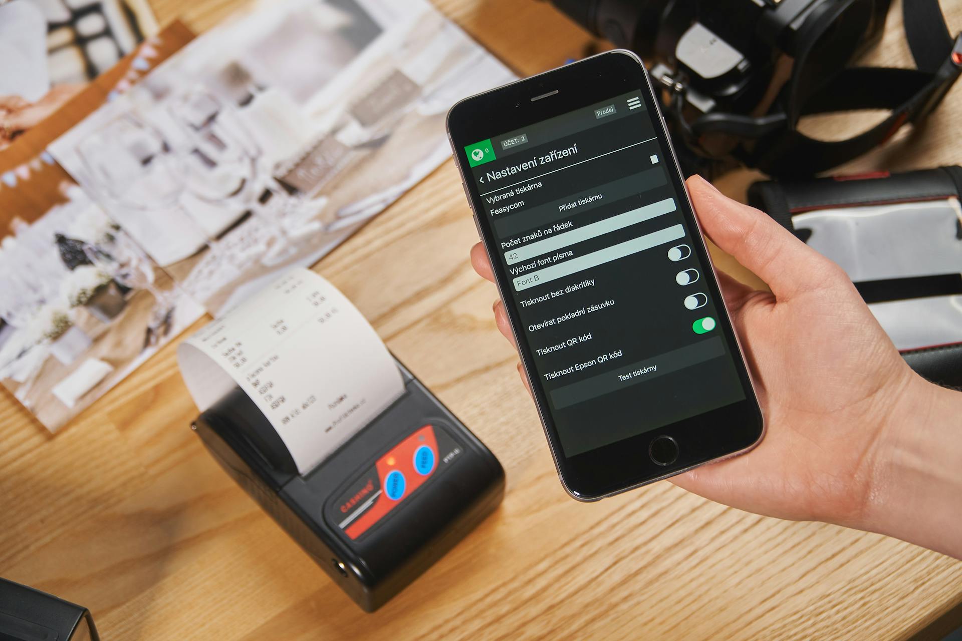 Close-up of a hand holding a smartphone with POS settings displayed, next to a receipt printer.