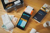 A modern payment terminal with receipts, cash, and manual on a wooden table.