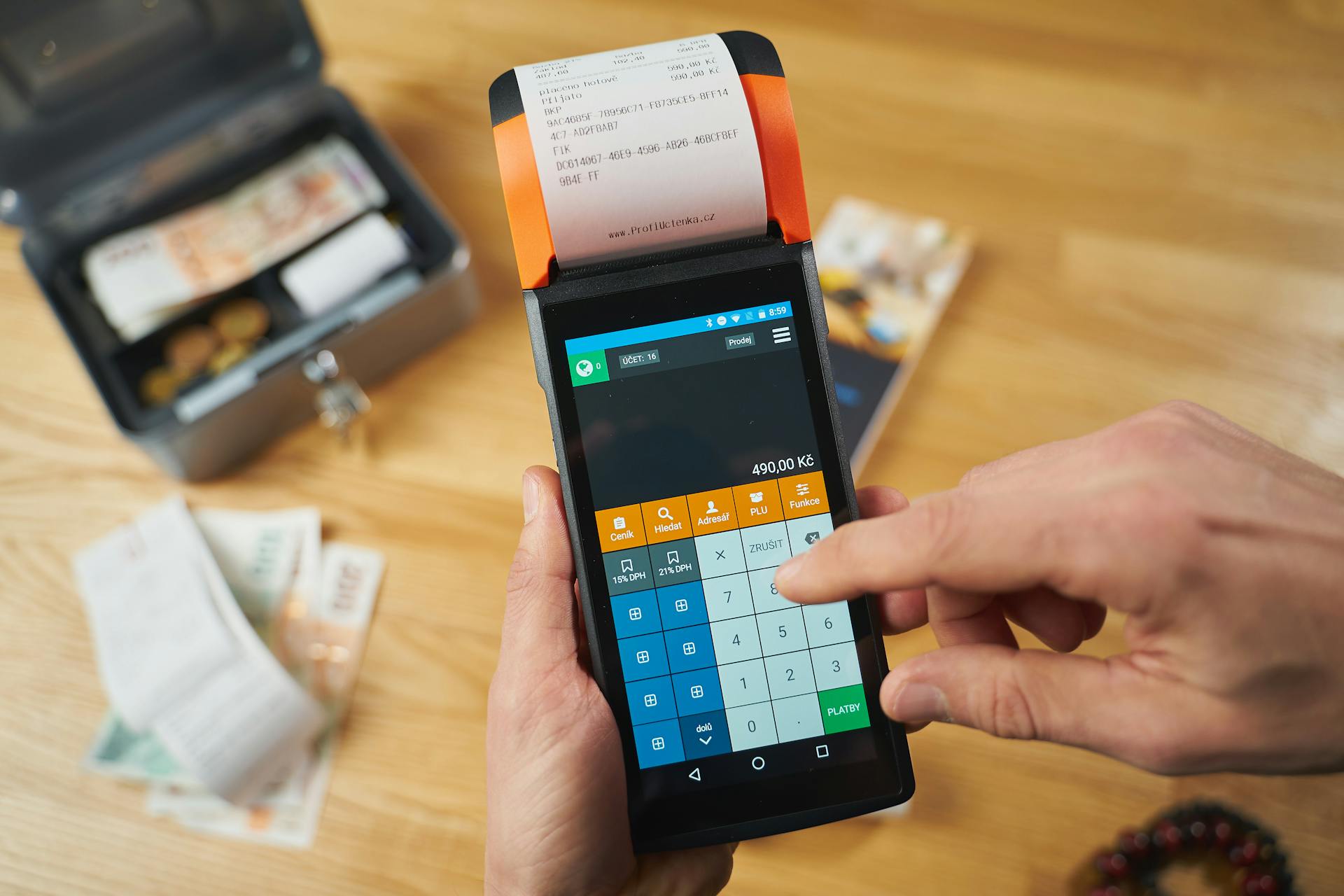 A close-up of a hand using a payment terminal with currency notes and a money box in view.