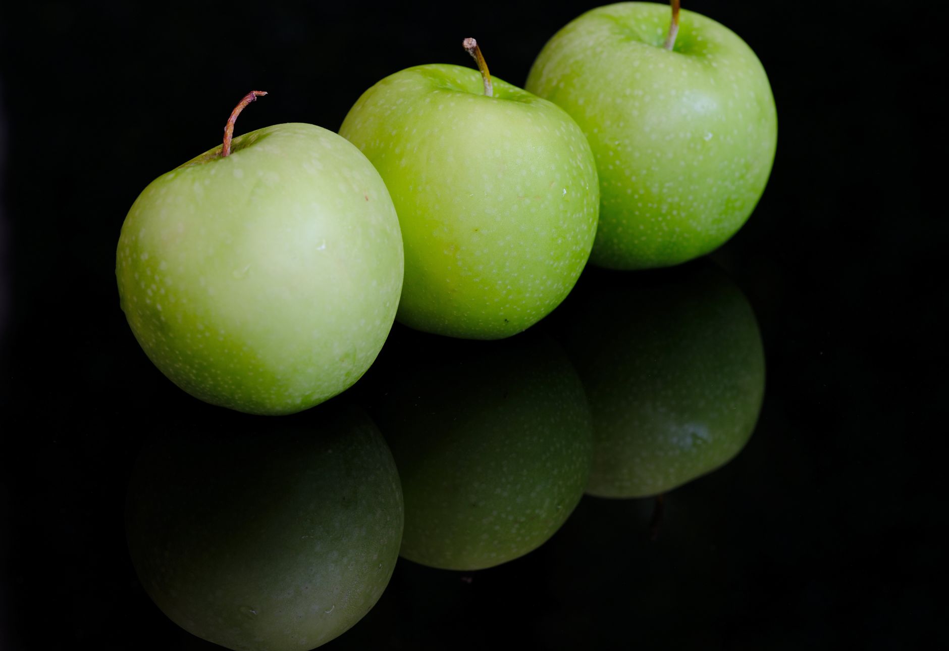Close-up of three green apples reflecting on a glossy black surface, showcasing freshness.