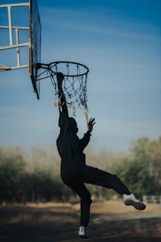 A person in silhouette performing a slam dunk on an outdoor basketball hoop during the day.