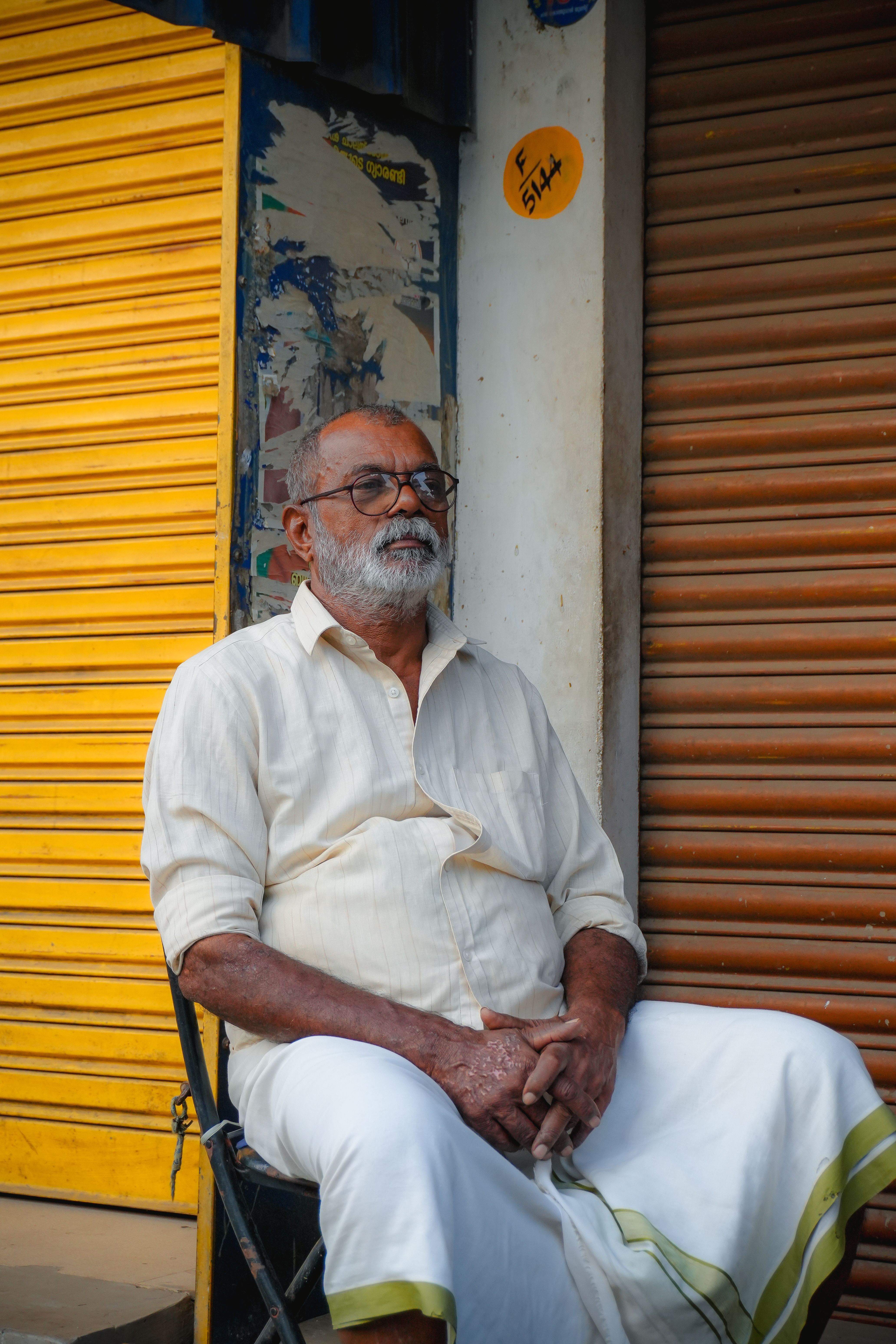 Elderly man in traditional attire sitting outside colorful shuttered shops.
